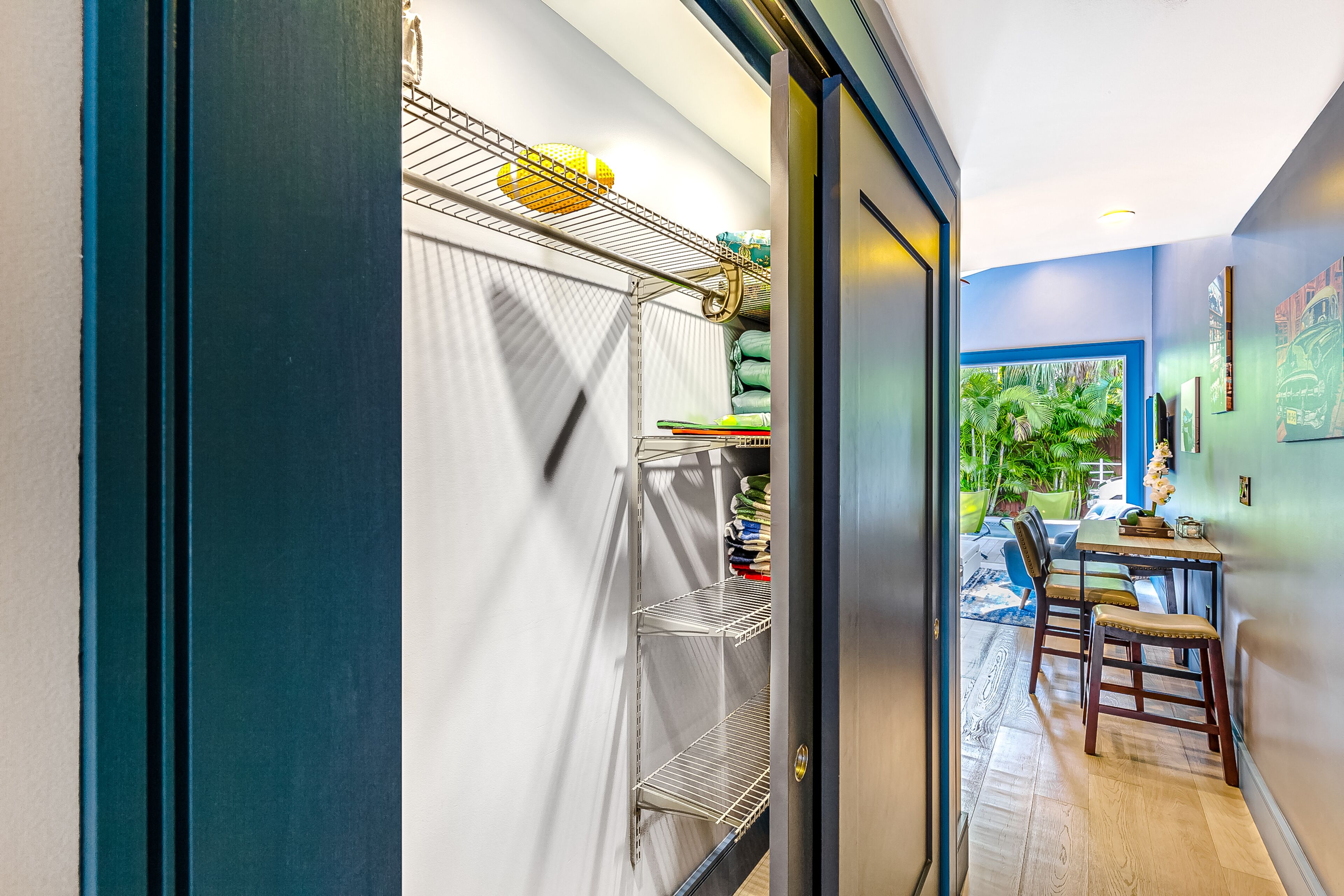 Un pasillo con una estantería metálica, una zona de comedor con mesa y sillas, y vistas de un jardín a través de un ventanal.