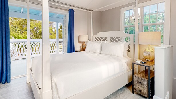 Uma cama grande com roupa de cama branca, uma mesa de cabeceira com abajur e vista para o verde lá fora.