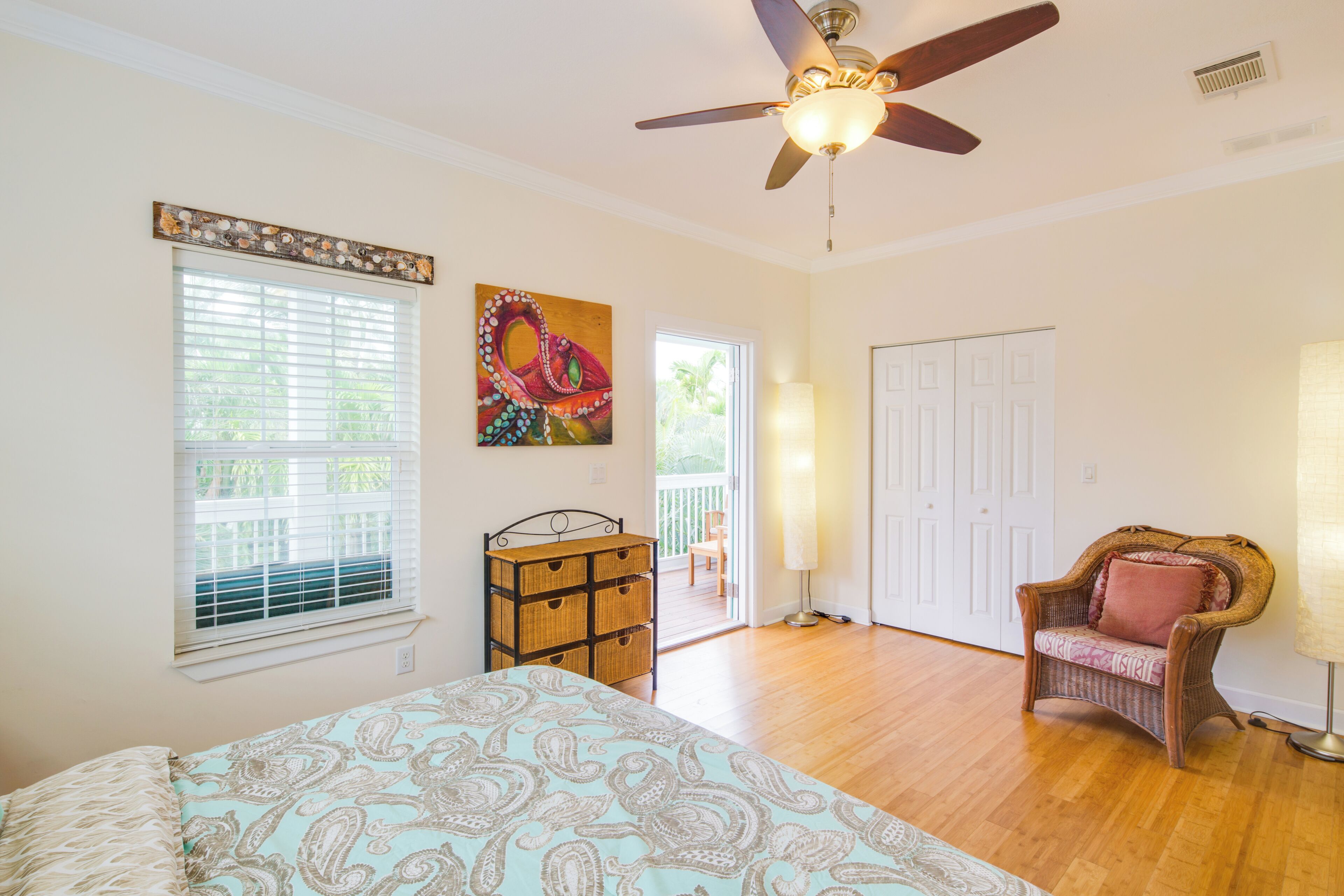 A bedroom with a bed, a ceiling fan, a window with blinds, a wicker chair, a door, and a painting on the wall.