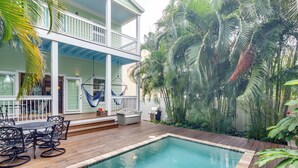 Uma área de piscina com deck de madeira, móveis de exterior e um edifício de dois andares com varanda.