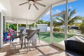 A covered patio area with wicker furniture, large windows, and a view of a green lawn and palm trees. - Quiet, spacious canal-side home with private pool, waterfront patio & central AC (Marathon)