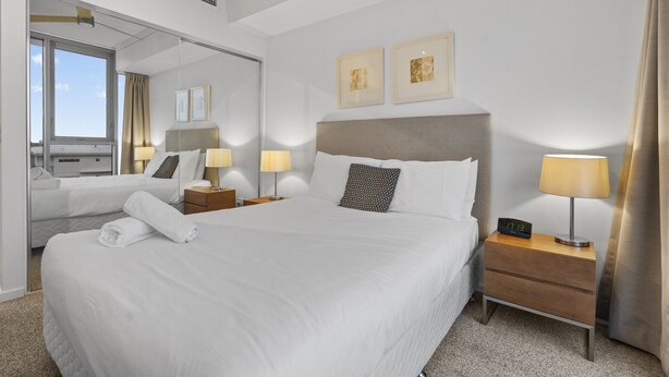 Quarto de hotel com uma cama grande, duas mesas de cabeceira com luminárias, um espelho e duas quadros na parede.