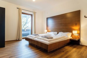 A double bed with a wooden headboard, two bedside tables with lamps, a large window with curtains, and a wooden floor. - Chalet Gretl by we rent (Zell am See)