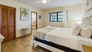Quarto de hotel com cama, ventoinha no teto, janela com cortinas, porta de madeira e uma pequena mesa.
