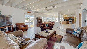 Ein großzügiger Wohnraum mit Ledersofas, einem Glastisch und einem Flachbildfernseher. Im Hintergrund ist eine Küche erkennbar, und der Raum verfügt über große Fenster.