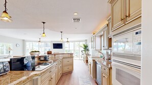A spacious kitchen with granite countertops, beige cabinetry, and a large island. There are pendant lights, a built-in oven, and a window providing natural light. - Oceanfront, Snowbird-Friendly Home with High-Speed WiFi, Ocean Views, and Dock (Key Largo)