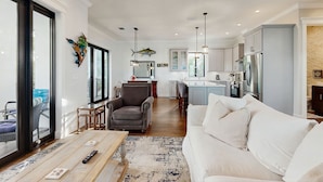 Een ruime woonkamer met een witte bank, een salontafel en een vloerkleed. In de achtergrond is een keuken met moderne apparatuur en een eethoek met stoelen te zien.