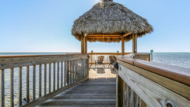 Une terrasse en bois mène à une structure couverte d’un toit de chaume, où se trouvent des chaises et une table, le tout offrant une vue sur la mer.