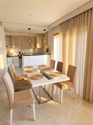 A modern dining area with a white table, beige chairs, woven placemats, and a kitchen in the background. - Casa Betz Alvor - 2 Bedrooms Apartment | Pool | Near Beach | Indoor Garage (Alvor)