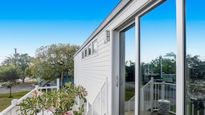 Une maison blanche avec une porte-fenêtre coulissante, une terrasse en bois et un petit jardin agrémenté de fleurs roses.