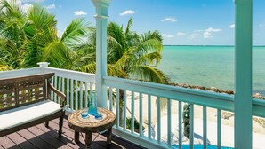 Una terraza de madera con un banco y una mesa, con vista a una playa de aguas turquesas cristalinas y palmeras.