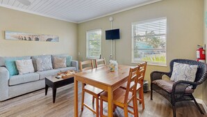 Un salon avec un canapé, une table à manger en bois et une chaise en osier. Il y a un tableau au mur et un téléviseur fixé au mur.