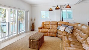 A spacious living room with a large tan leather sofa, a wicker ottoman, and two pendant lights. - Waterfront Half-Duplex with 30-Ft. Dock, Sitting Pool, and Private Beach Access (Key Colony Beach)