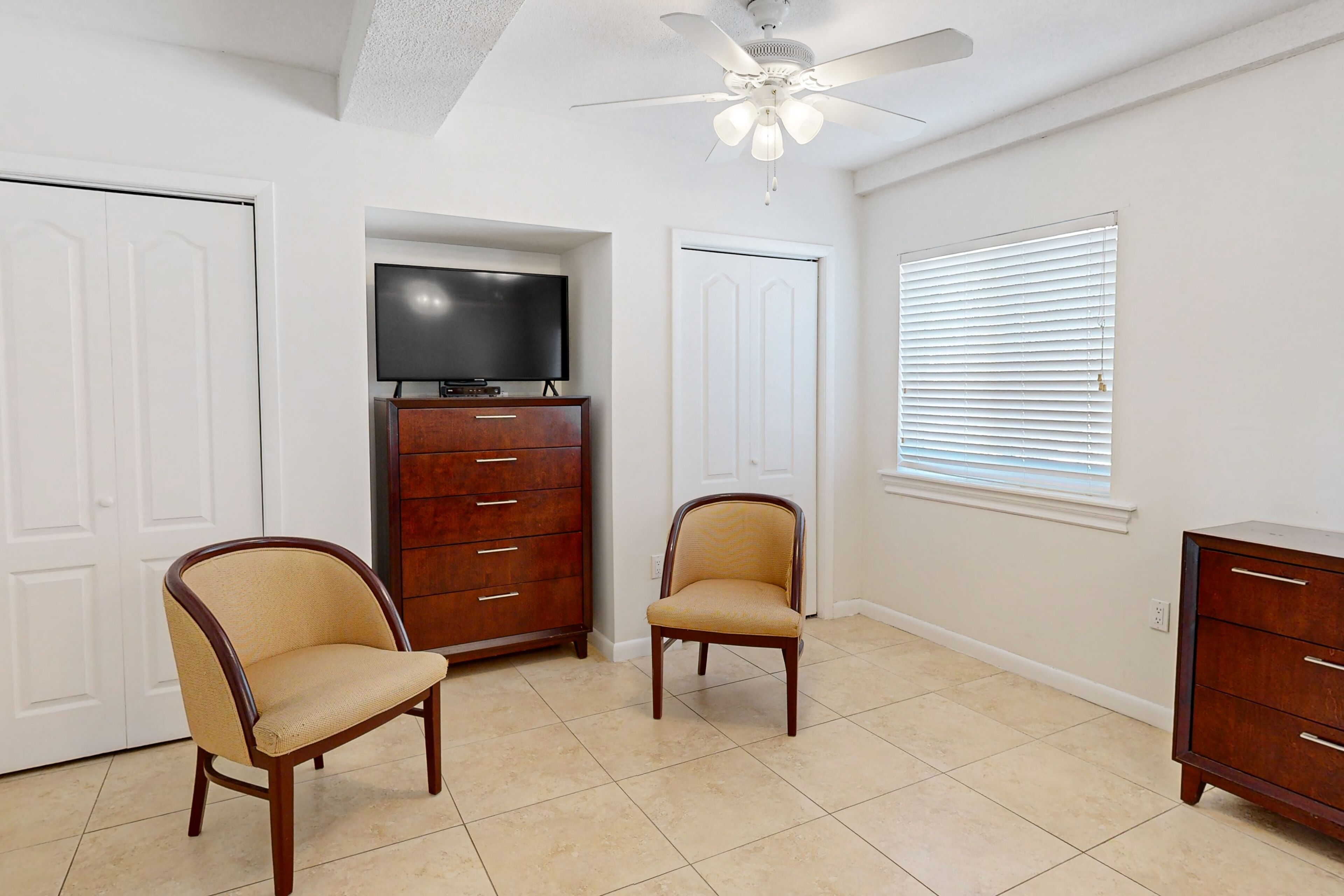A room with two wooden chairs, a television on a wooden cabinet, a ceiling fan, and a window with blinds.