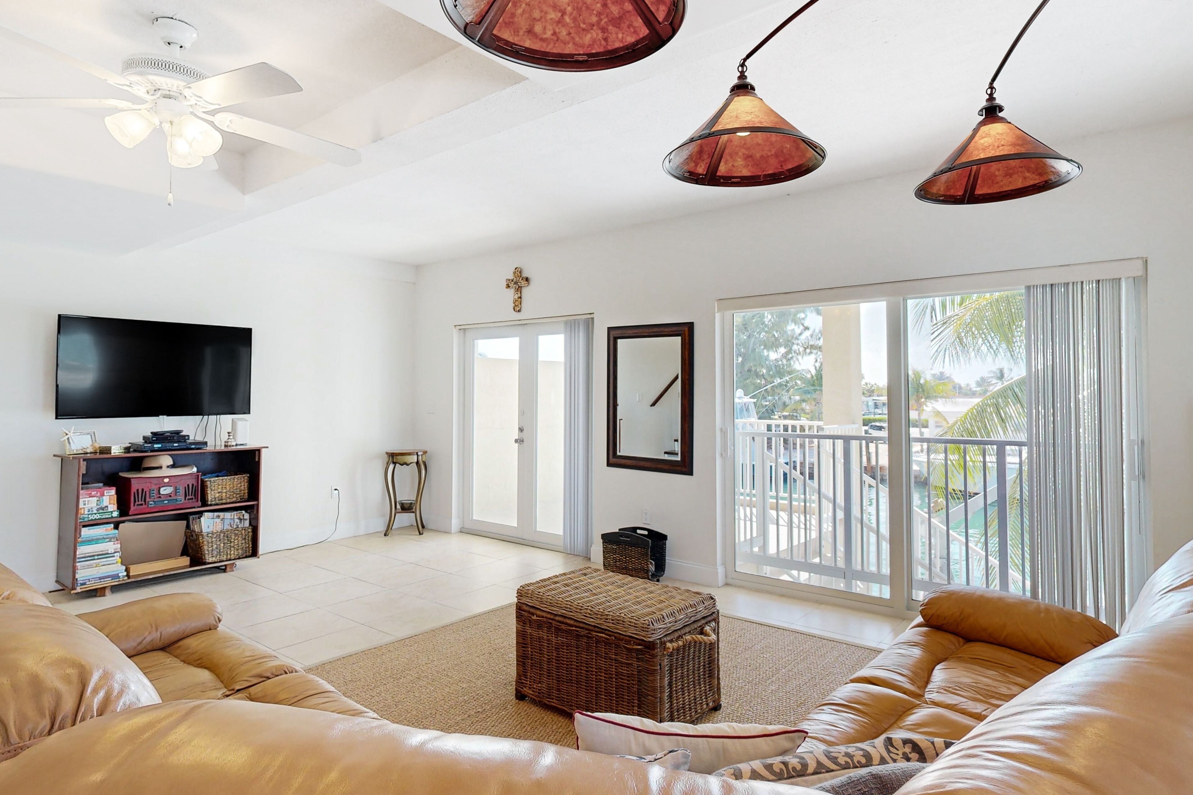 A living room with a large flat-screen TV, wicker ottoman, leather sofas, and a ceiling fan.