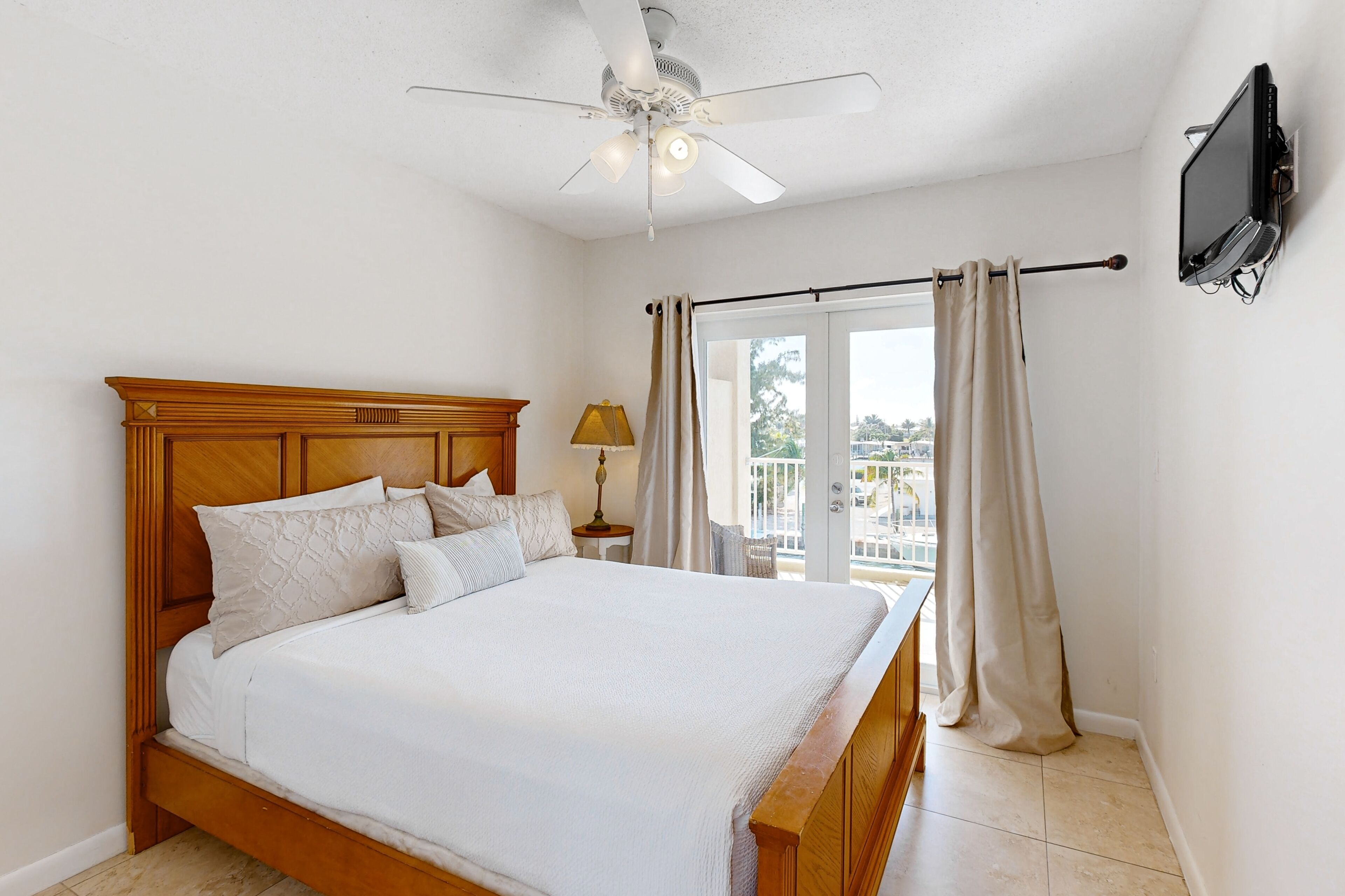 A bedroom with a wooden bed, a ceiling fan, a TV mounted on the wall, and a view of the outdoors through a sliding door.