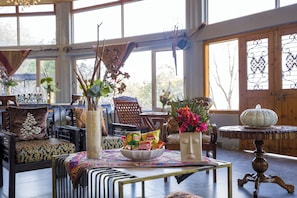 A room with a large window, a wooden door, a table with a bowl of chips, and various chairs and tables. - ELIVAAS Zephyr Sierra 2-BHK Hillside (Dhanaulti)