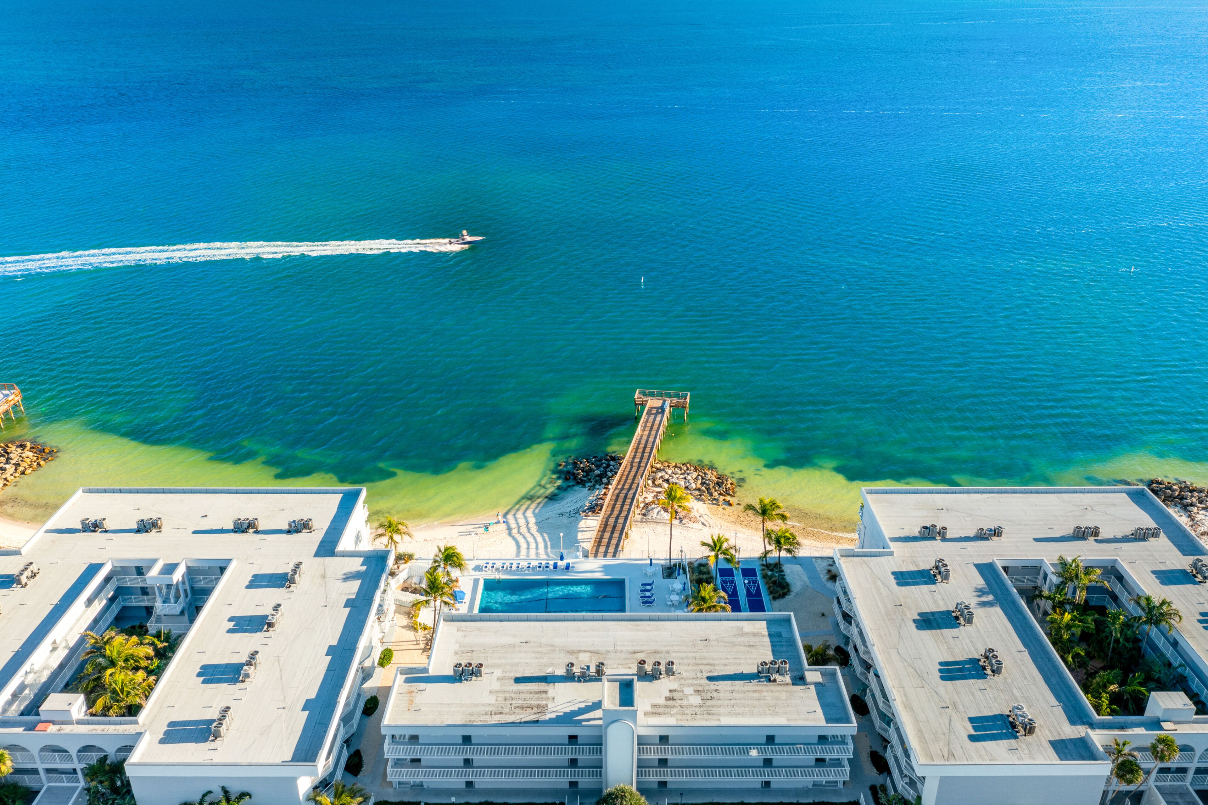 An aerial view of a hotel with a rooftop pool, a pier extending into the sea, and a beach area with palm trees.