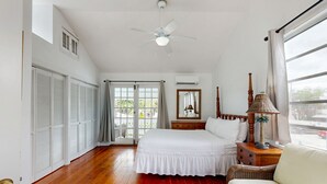 Un dormitorio con cama, ventilador de techo, ventana con cortinas, mesita de noche y una silla de mimbre.