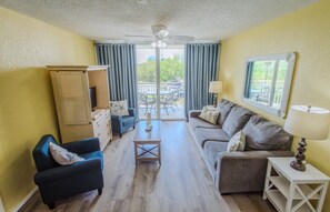 Un salon avec un ventilateur de plafond, un canapé, un fauteuil, une petite table et une fenêtre avec des rideaux.