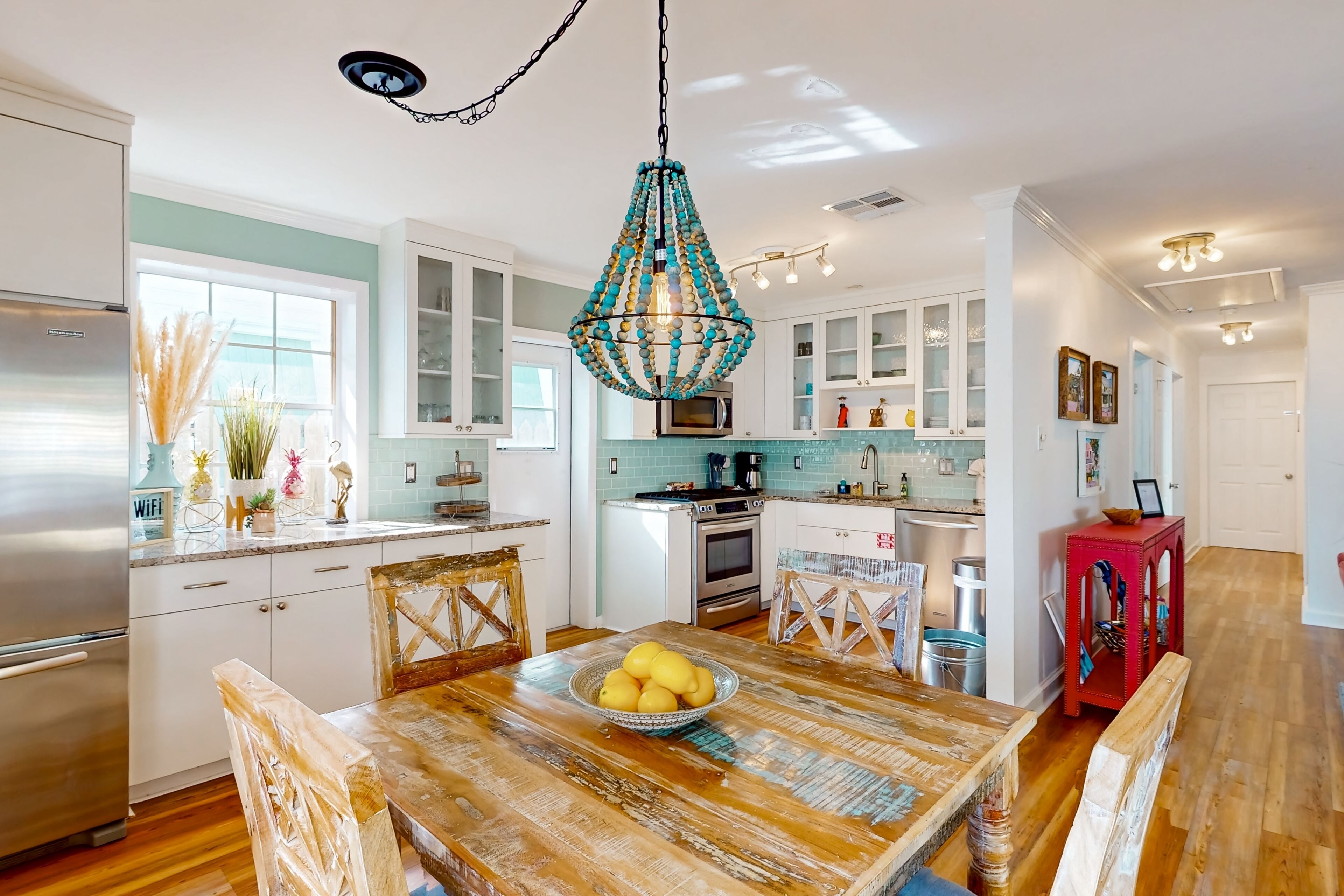 A modern kitchen with white cabinets, a wooden dining table with a bowl of lemons, and a decorative chandelier.