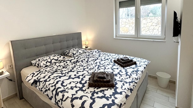 A bedroom with a bed, a nightstand, a window, and a television mounted on the wall. - Apartment Mariner 1 With Heated Pool (Nin)