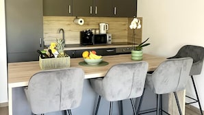 A modern kitchen with dark cabinets, a wooden countertop, a coffee machine, and a bowl of fruit. There are three grey chairs around the table. - Apartment Mariner 1 With Heated Pool (Nin)