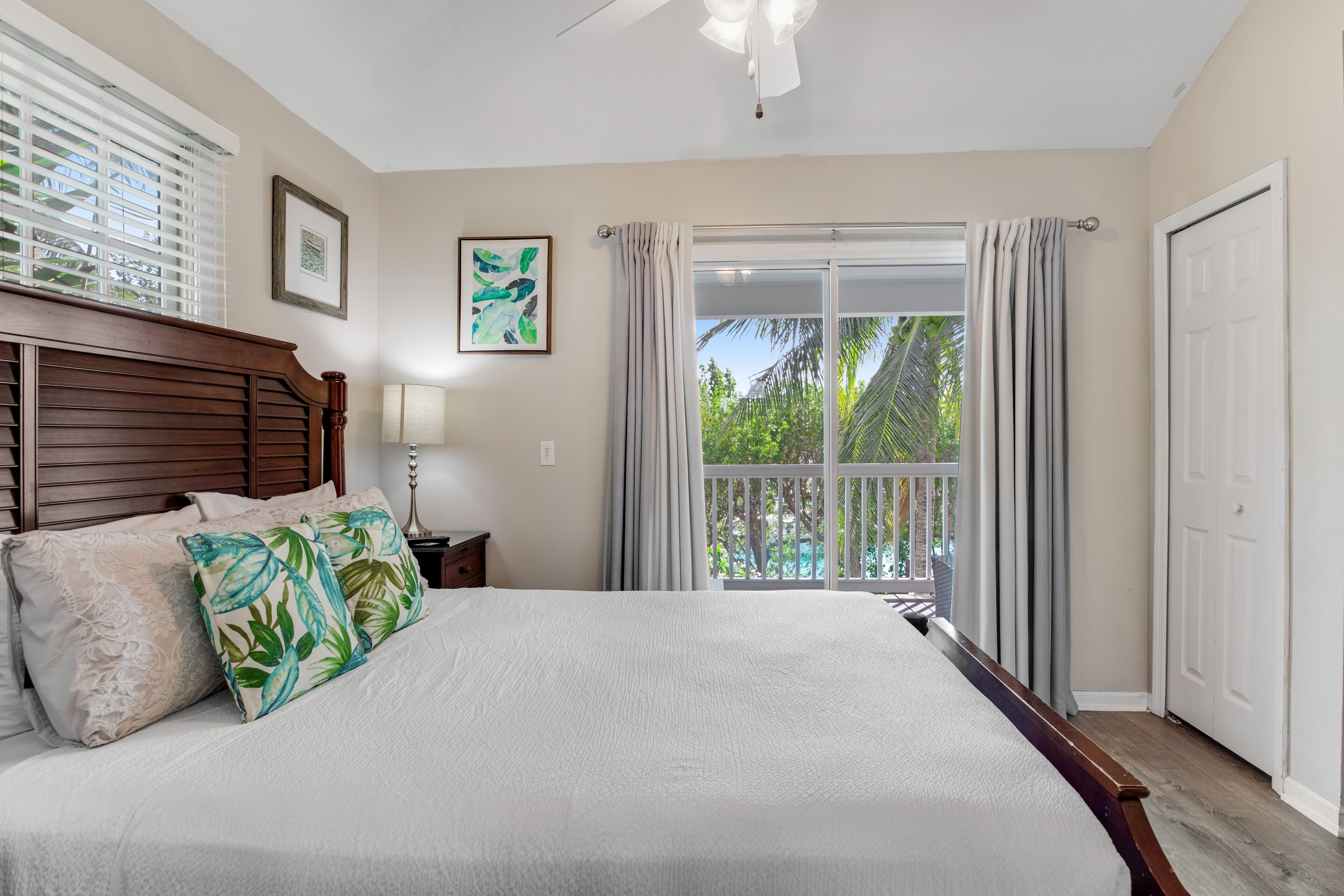 A bedroom with a large bed, a nightstand, a ceiling fan, a window with blinds, and a view of greenery outside.