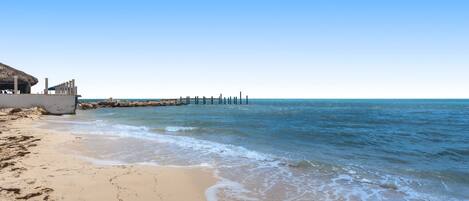 A beach with a thatched-roof structure, a wooden pier, and clear blue skies.