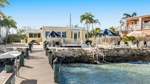 A waterfront property with a wooden deck, clear blue water, and a small building with a balcony. - Waterfront beauty with a dock, small private beach, washer/dryer, & gas grill (Key Colony Beach)