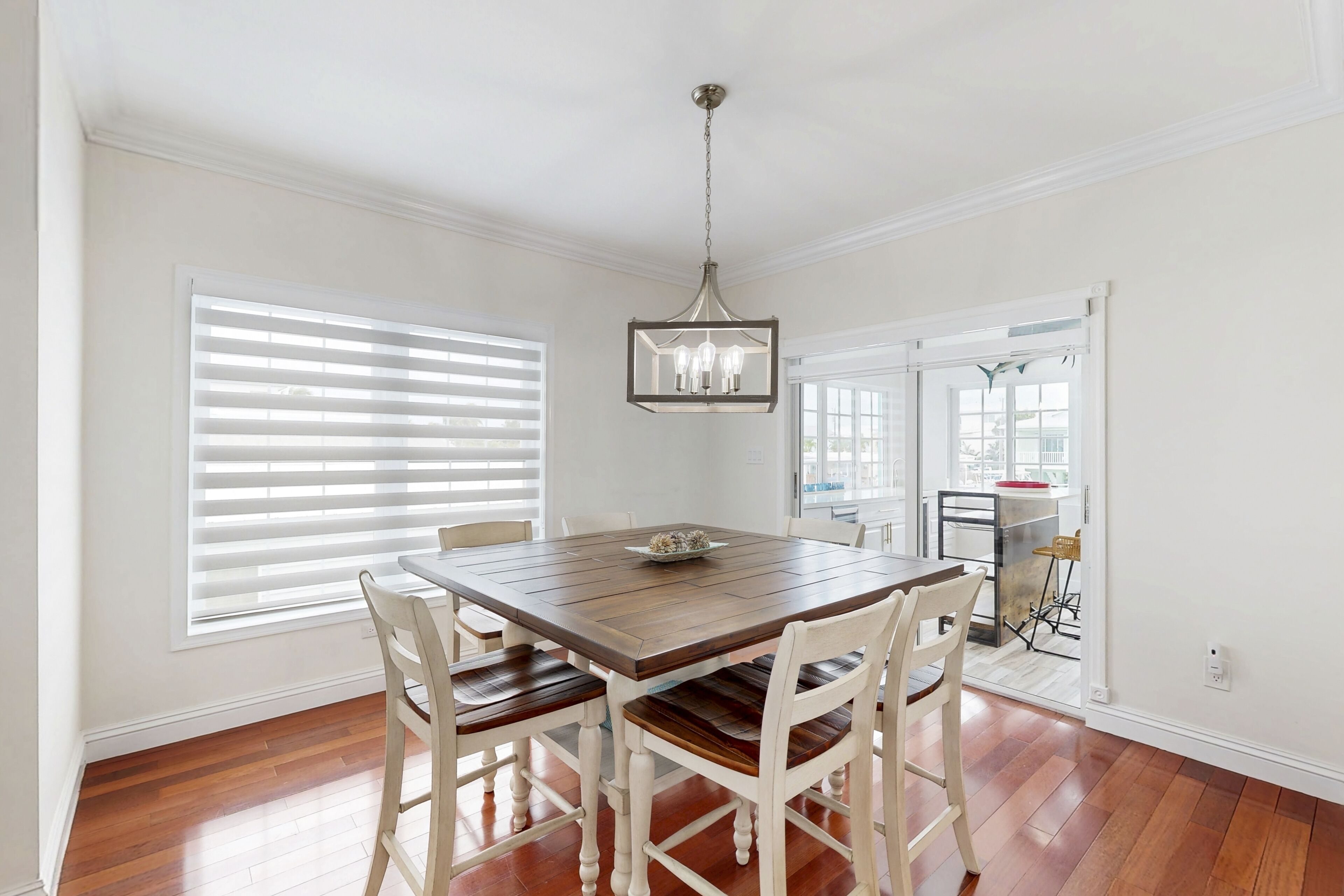 A dining area with a wooden table and white chairs, a hanging light fixture, and large windows with blinds.