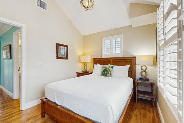 A bedroom with a large bed, wooden headboard, two bedside tables with lamps, a window with blinds, and a door leading to another room. - Tropical oasis near all the action with central AC & balcony (Duck Key)