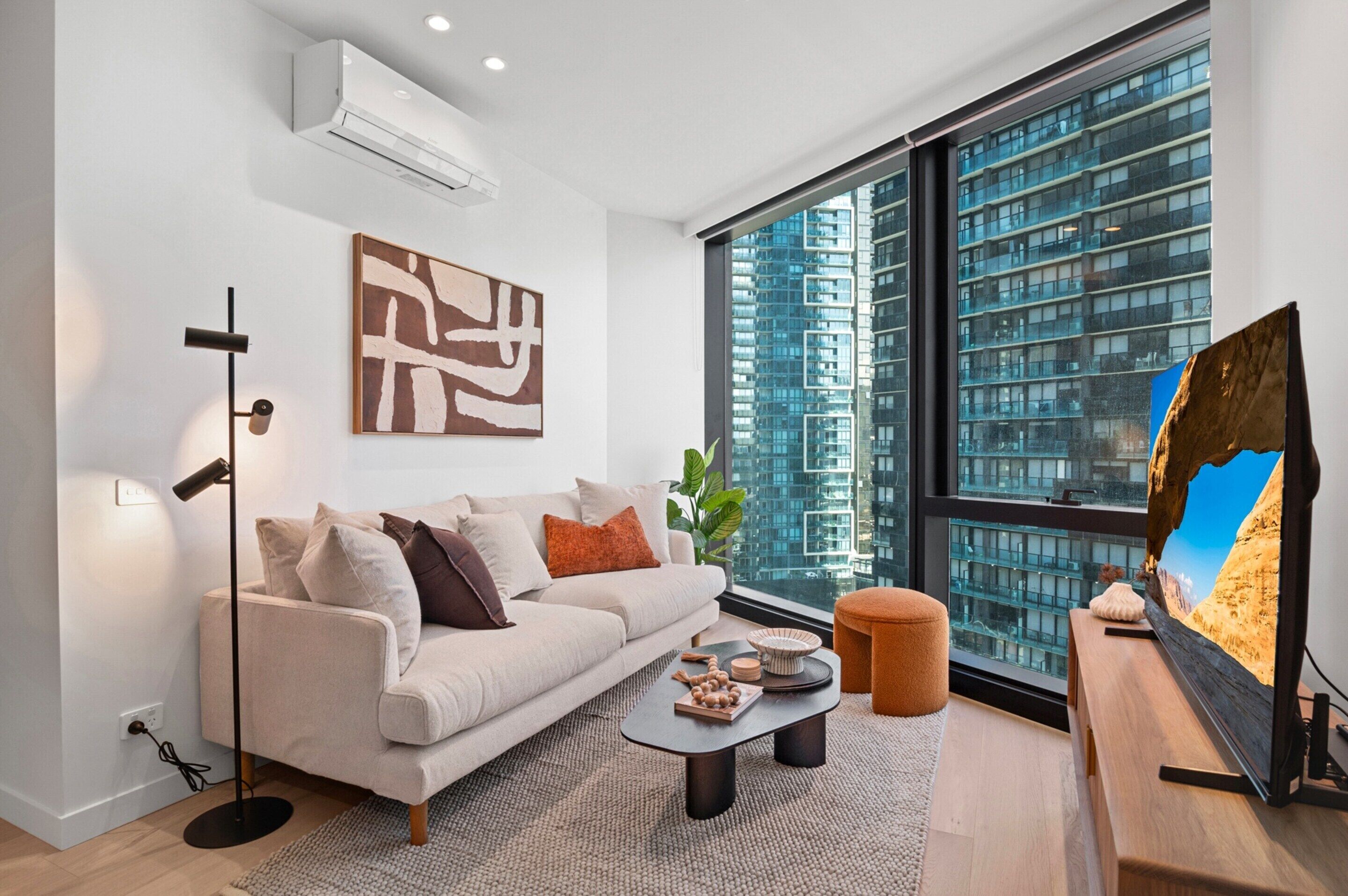 Un salón moderno con sofá, mesa de centro y obras de arte, ventanas amplias con vista a la ciudad y televisor de pared.