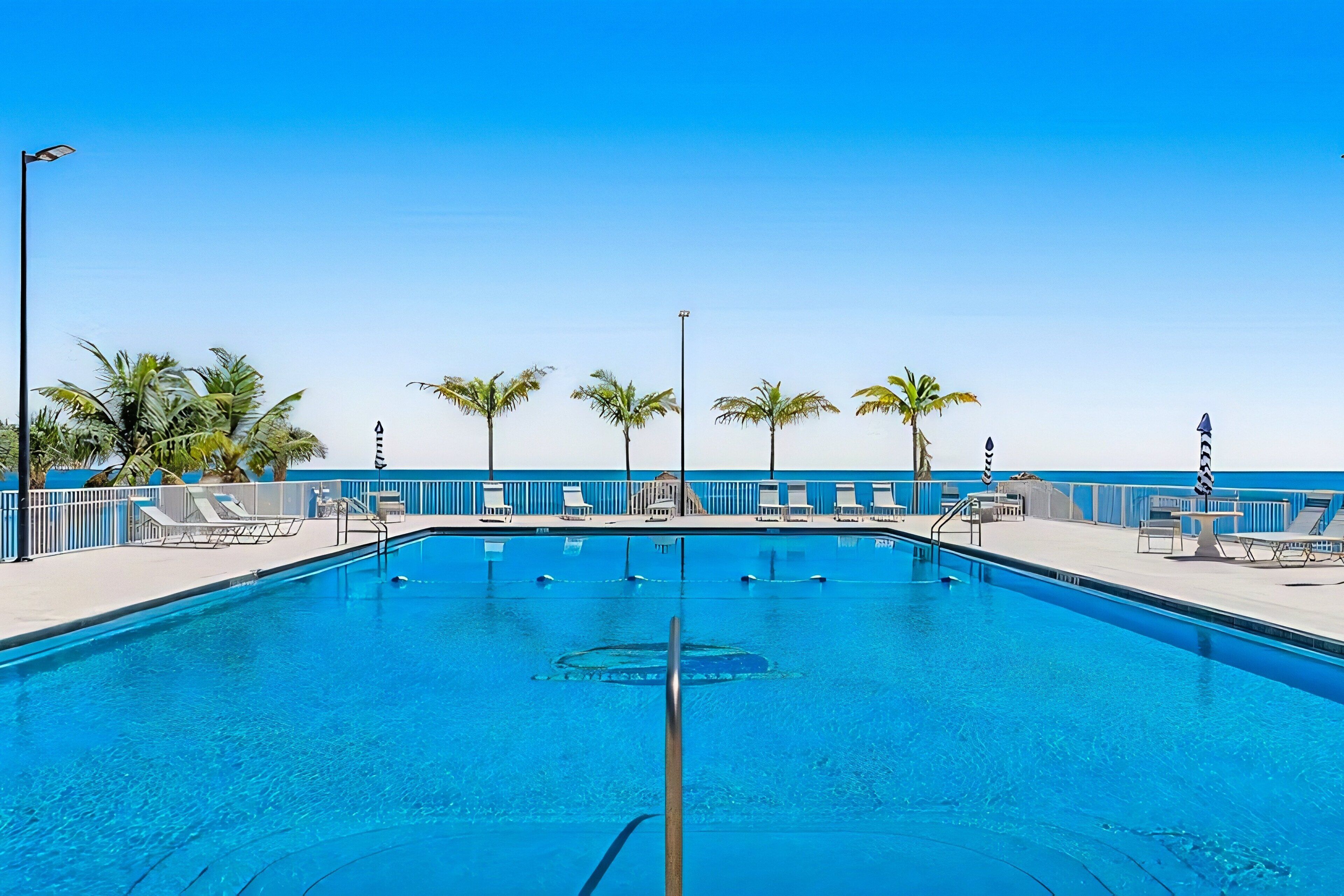 A clear blue pool with lounge chairs, palm trees, and a view of the ocean.
