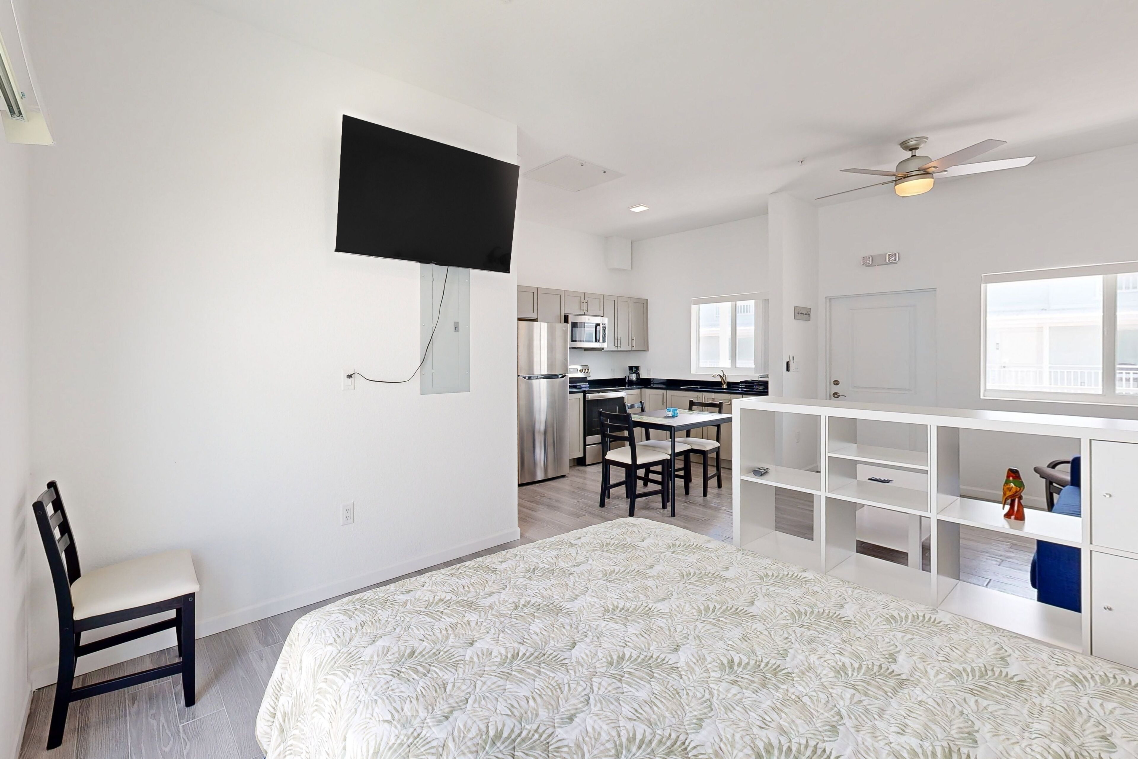 Une chambre moderne avec un grand lit, un bureau et une chaise. On aperçoit au fond une petite cuisine équipée d’un four à micro-ondes et d’un réfrigérateur.