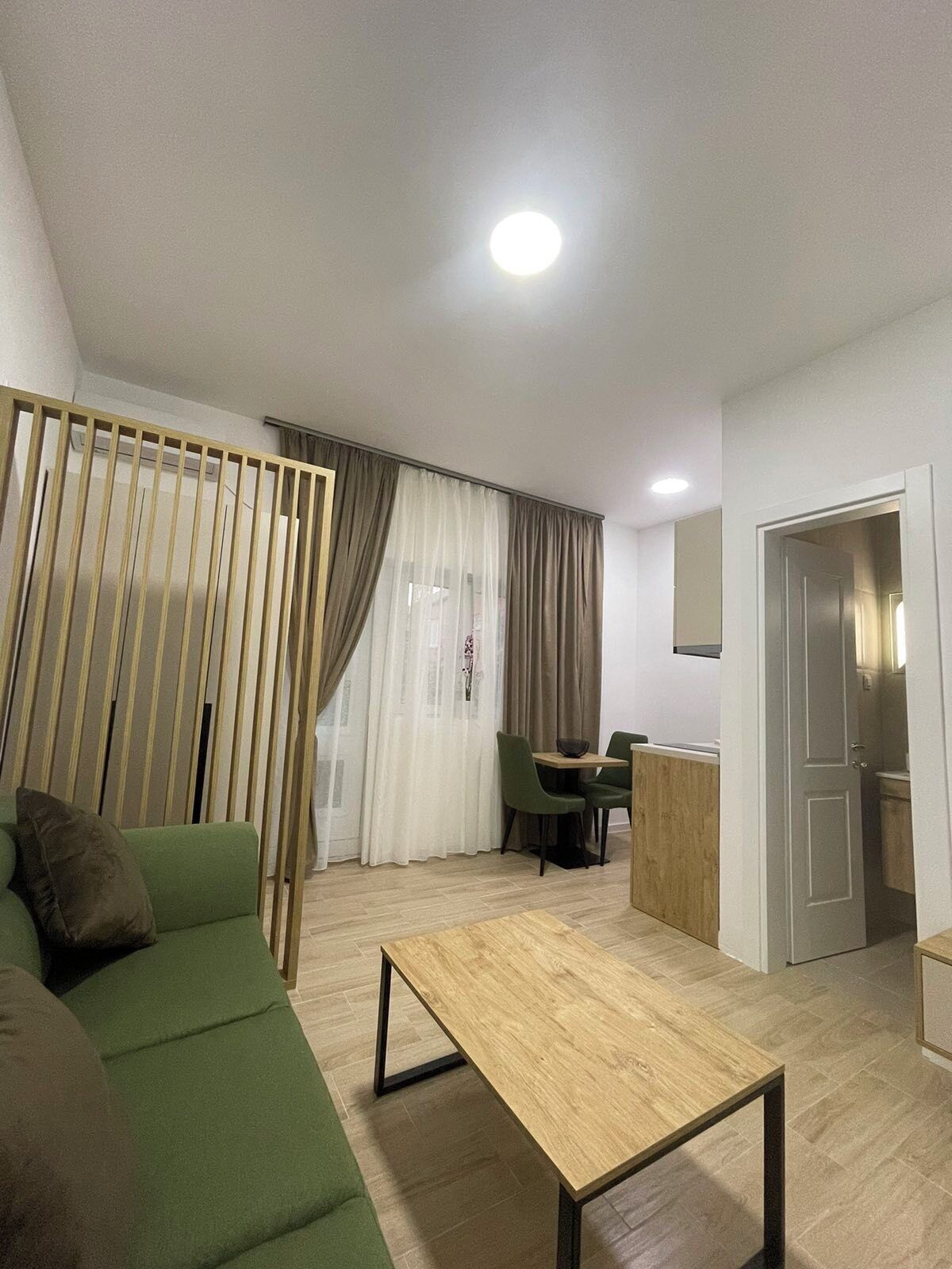 A modern living room with a green sofa, a wooden coffee table, a dining area with a table and chairs, and a kitchen area with a cabinet.