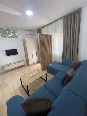 Una sala de estar moderna con un sofá azul, una mesa de centro de madera, un televisor en la pared y una ventana con cortinas.