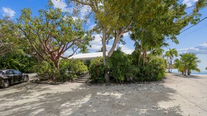 Une propriété située en bord de mer, avec une voiture garée sur le sable, un bâtiment blanc et une végétation luxuriante.