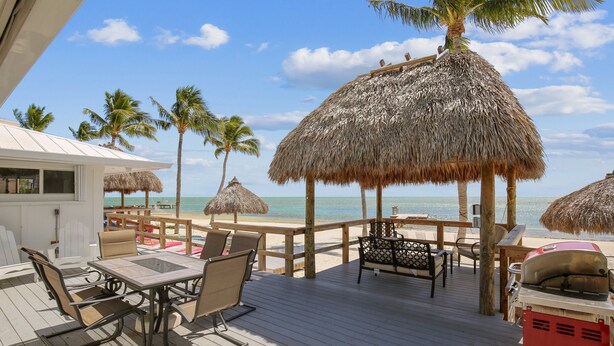 Une terrasse en bois agrémentée de parasols en chaume, de chaises de plage et d’un barbecue.