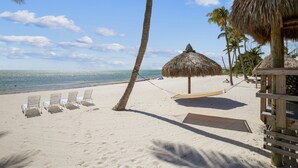 Une plage avec des chaises longues blanches, un hamac et une structure recouverte d’un toit de chaume.