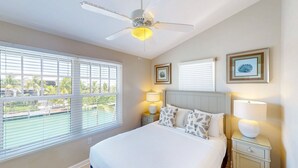 A bedroom with a bed, ceiling fan, bedside lamps, and a window with blinds. - Enchanting waterfront townhouse with balcony, mini oasis pool, & washer/dryer (Duck Key)