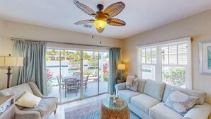 A living room with a ceiling fan, a sofa, a chair, a glass coffee table, and a view of an outdoor patio with a pool and palm trees. - Enchanting waterfront townhouse with balcony, mini oasis pool, & washer/dryer (Duck Key)