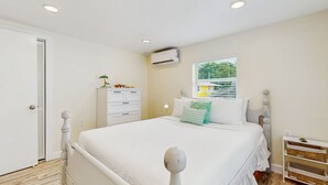Ein Schlafzimmer mit einem weißen Bett, einem Schminktisch, einem Fenster mit Jalousien und einer Klimaanlage.