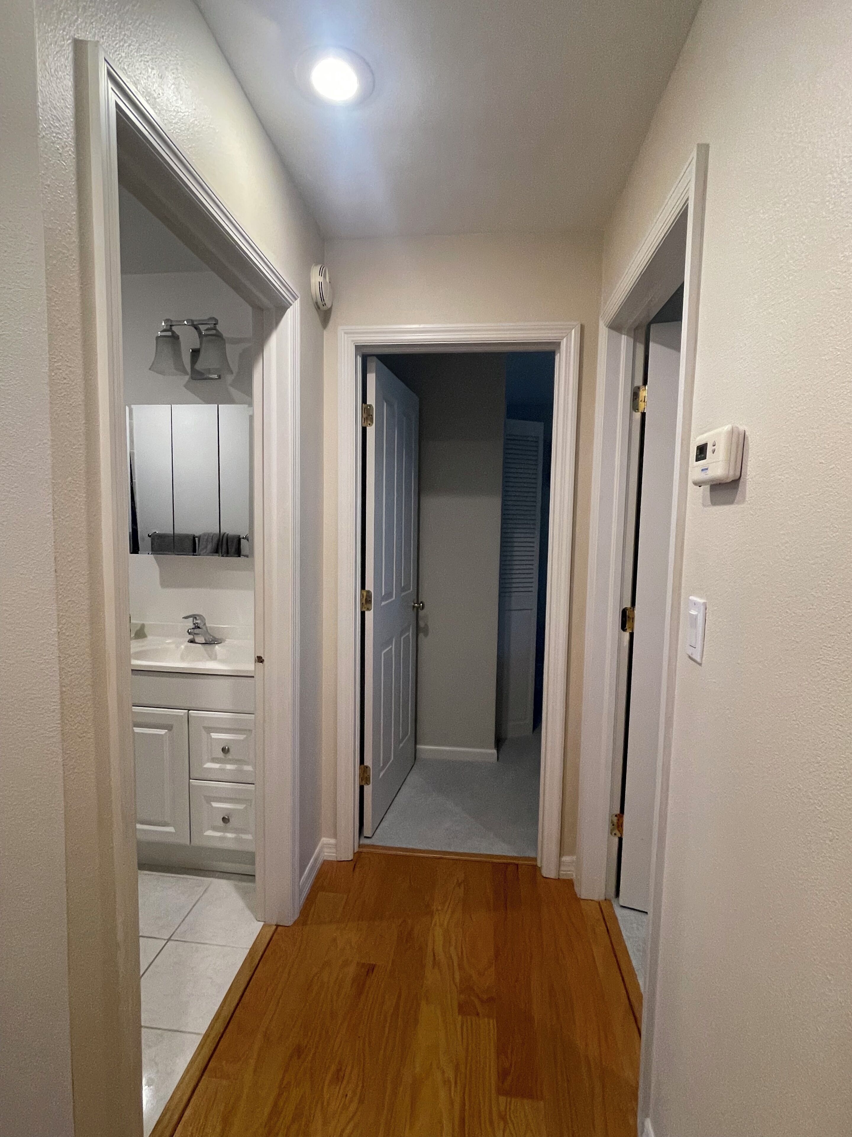 A hallway with wooden flooring, white walls, and a door leading to a bathroom with a sink and mirror.