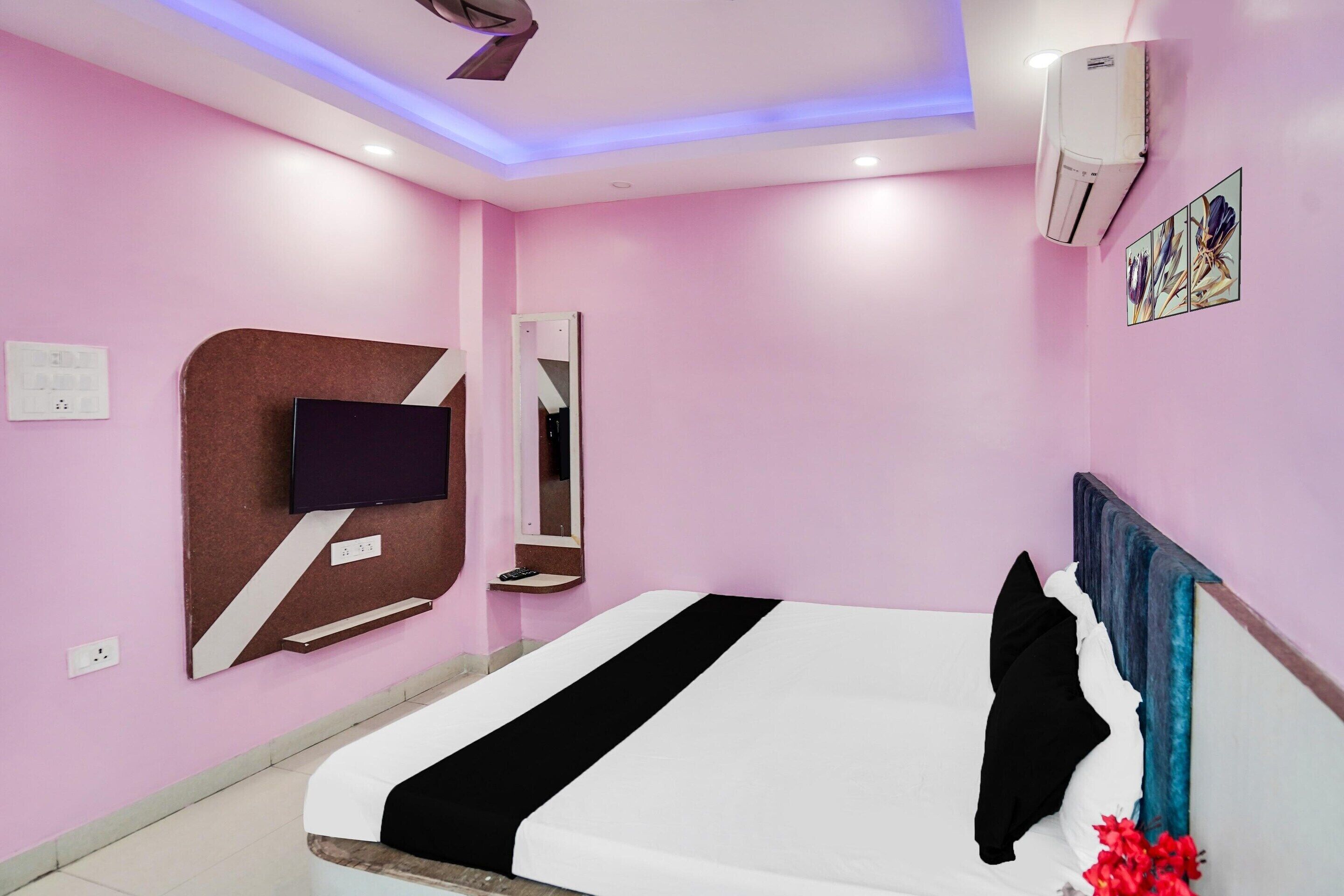 A pink-walled hotel room with a bed, a mounted TV, a mirror, and an air conditioning unit.