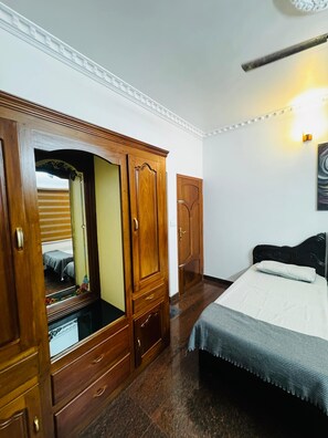 Un dormitorio con un armario de madera, una cama con colchón blanco y cabecera oscura, y una lámpara fijada a la pared.