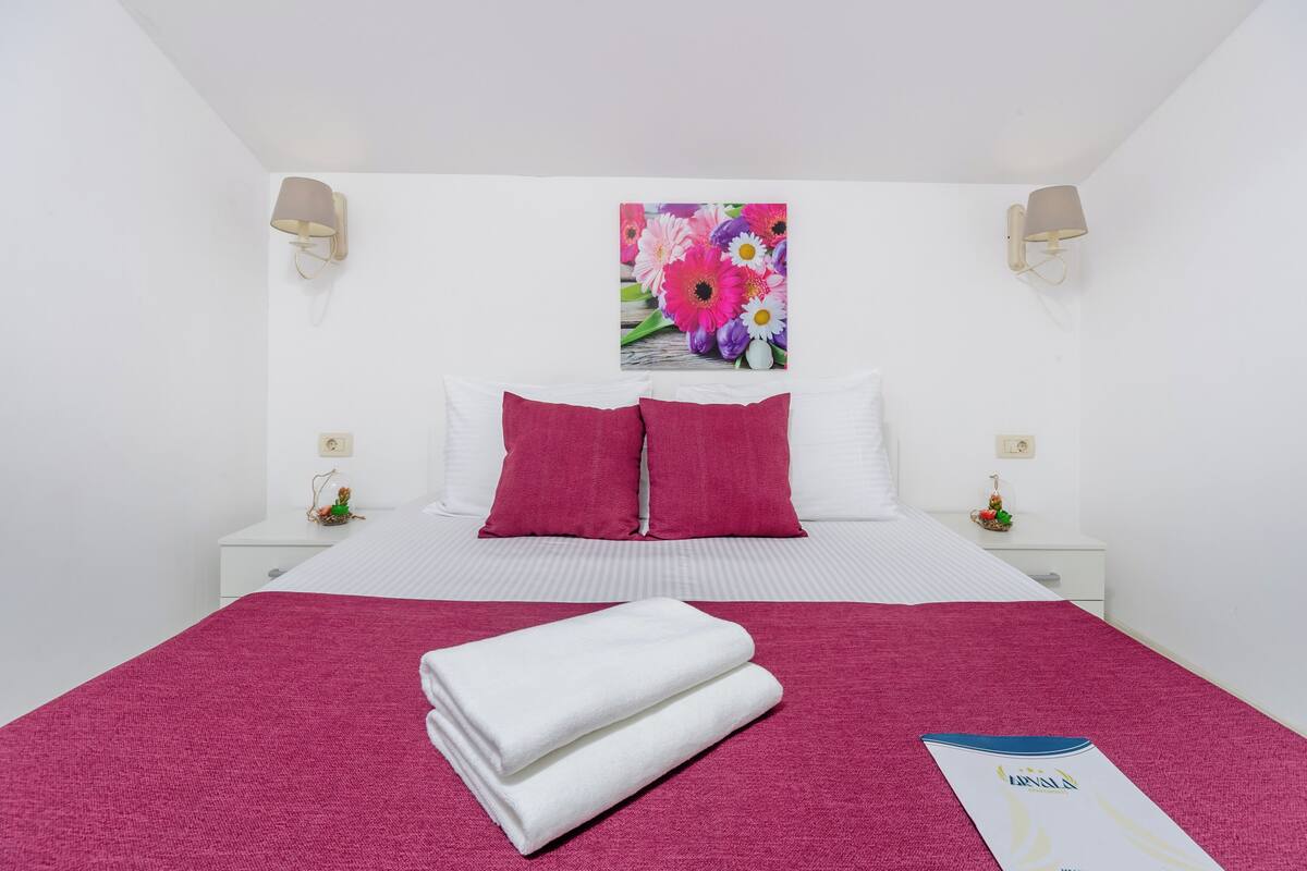 Ein Hotelzimmer mit Bett, Kissen, einem Nachttisch, einer Wandlampe und einem gerahmten Blumenbild.