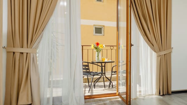 Una habitación con puerta corredera de vidrio que da a un balcón con mesa y sillas, lámpara colgante y vista de un edificio.