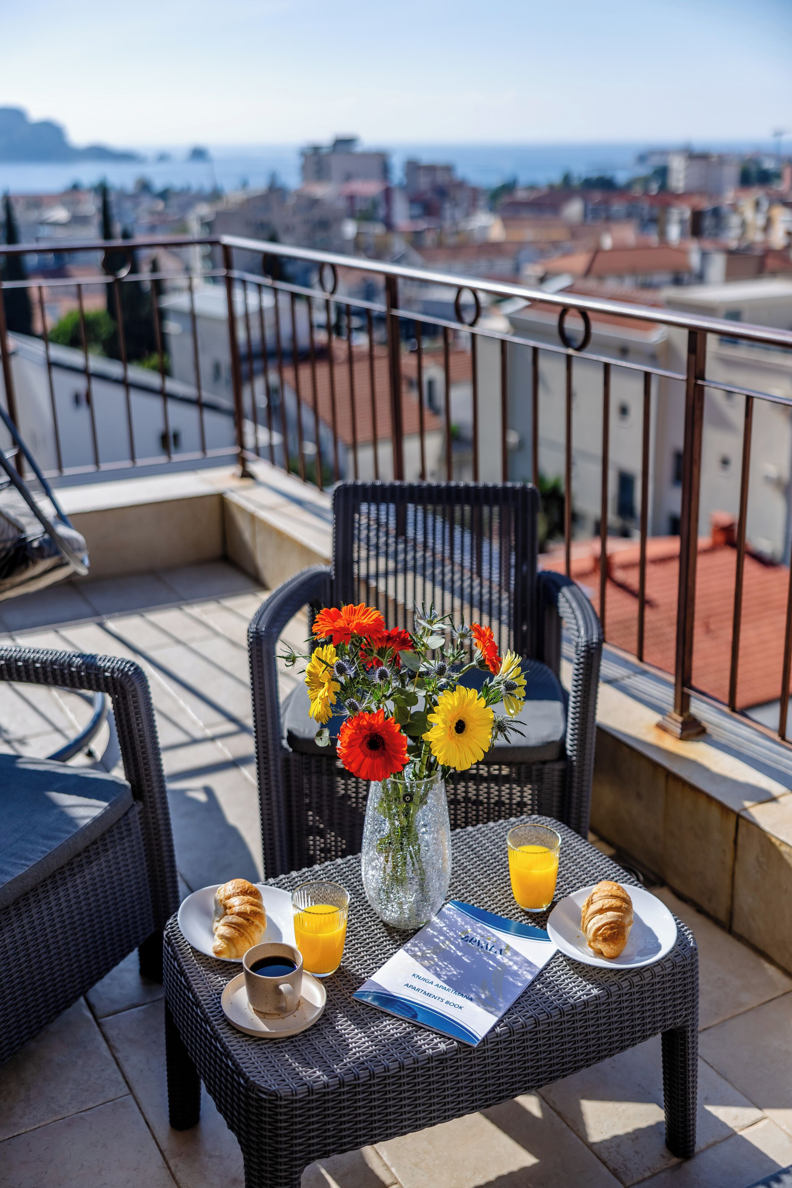 Un balcón con mobiliario de mimbre, un florero con flores, croissants, un vaso de jugo de naranja y un menú.