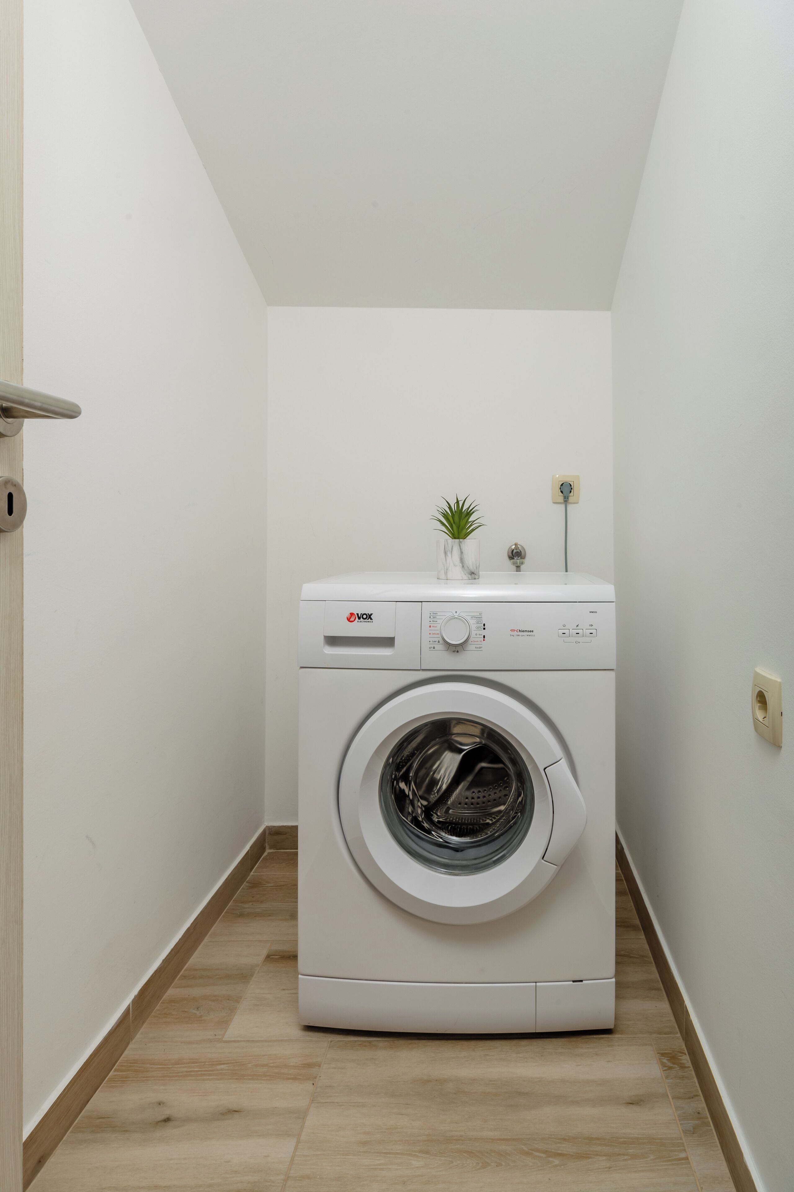 A compact, white, wall-mounted washing machine in a narrow hallway with a wooden floor, a small plant on top, and a power outlet on the wall.